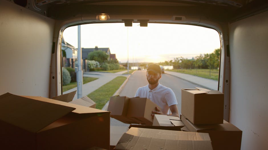 A person wearing a navy blue vest, black trousers, and a white cap is standing at the rear of a white moving van with open double doors, which reveals an interior filled with neatly stacked cardboard