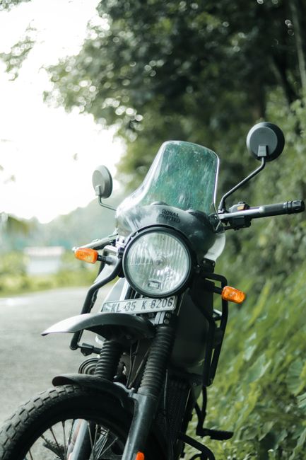 A black motorcycle equipped with a large, transparent windshield and rearview mirrors is parked on a paved road adjacent to a grassy area with dense green foliage and tall trees in the background. The motorcycle features a round headlight, orange turn signals, and a front wheel with a visible tire. The background shows a blurred view of greenery and the sky, indicating a daytime outdoor setting. The scene is associated with house removals and moving logistics, as part of the transportation process facilitated by Enfield Movers, a company specializing in removals in the Enfield area, focusing on packing, loading, and furniture transport services.
