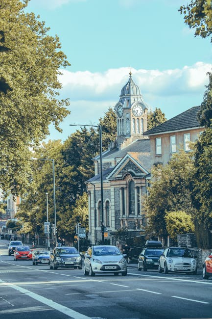 A street view in Enfield Town EN1 featuring a historic church with a tall clock tower topped by a spire, surrounded by leafy trees and a clear blue sky. In the foreground, several parked cars are situated along the curb while a few moving vehicles are visible on the road. The scene captures a typical daytime setting with natural sunlight illuminating the buildings and street, which is part of a home relocation or furniture transport scene. Occasionally, Enfield Movers' mobile units might be seen nearby during house removals or moving services, and the area includes pavement and street fixtures such as lampposts and traffic signs, supporting the context of local logistics and moving activities.