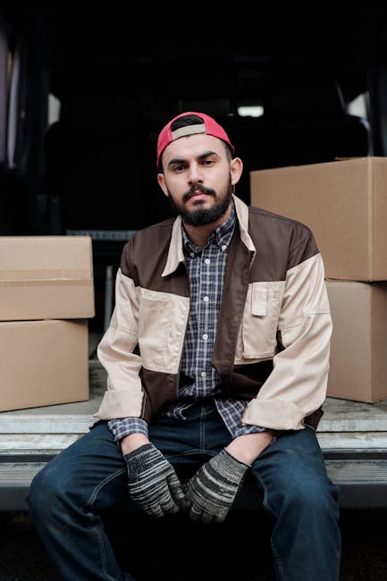 A young man with a beard, wearing a red cap backwards, a plaid shirt, and a beige and brown jacket, sitting on the edge of a loading area at the back of a van during a house removal process. Behind him inside the van are several cardboard boxes of various sizes, some stacked and some placed on the floor, prepared for packing and transportation. The van is parked on a driveway or curbside, with a dark interior visible in the background. The man appears to be taking a break during the furniture transport phase of a home relocation, with the boxes ready for loading or unloading. Enfield Movers’ professional removal service is involved in the secure packing and careful loading of items, which include cardboard packaging materials and the use of straps or blankets if visible, to ensure furniture and belongings are transported safely. The scene captures the practical aspects of the moving and packing process at a residential property.