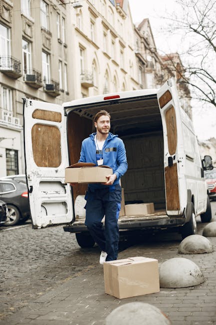 A young man with a beard, wearing a red cap backwards, a plaid shirt, and a beige and brown jacket, sitting on the edge of a loading area at the back of a van during a house removal process. Behind him inside the van are several cardboard boxes of various sizes, some stacked and some placed on the floor, prepared for packing and transportation. The van is parked on a driveway or curbside, with a dark interior visible in the background. The man appears to be taking a break during the furniture transport phase of a home relocation, with the boxes ready for loading or unloading. Enfield Movers’ professional removal service is involved in the secure packing and careful loading of items, which include cardboard packaging materials and the use of straps or blankets if visible, to ensure furniture and belongings are transported safely. The scene captures the practical aspects of the moving and packing process at a residential property.