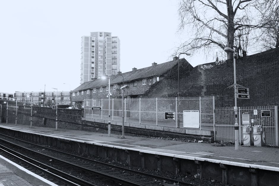 A black and white photograph of Ponders End railway station platform, showing railway tracks in the foreground with wooden sleepers and metal rails, bordered by a concrete edge. Along the platform, there are several metal lampposts with lights attached, illuminating the area. Behind the platform, a tall brick wall separates the station from residential buildings, which include a small, single-story brick house with a pitched roof and multiple chimneys, and a taller modern high-rise building in the background. The high-rise has a white exterior with numerous windows, contrasting with the older, darker brick structures. The platform appears empty, with no visible people or moving equipment, and there are signs and noticeboards attached to the fencing and walls. The scene depicts a typical urban railway station environment, with a focus on the infrastructure involved in home relocation, furniture transport, and packing and moving preparations, aligning with services provided by Enfield Movers.