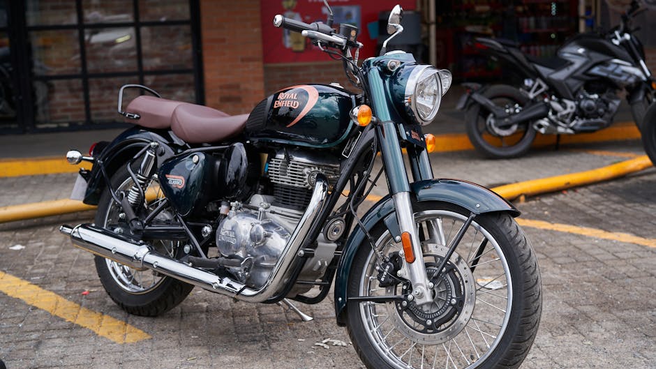A vintage Royal Enfield motorcycle parked in a designated parking area on a paved surface, with a brown leather seat, black fuel tank, and chrome exhaust pipe visible. The motorcycle features a round headlight, front fender, and spoked wheels, with orange turn signals mounted on the front. In the background, there are two other modern motorcycles parked parallel to the royal Enfield, with one partially visible, having a black body and silver accents. The parking area is bordered by a low yellow curb, and a brick building with large windows can be seen behind the motorcycles. The scene is illuminated by natural daylight, with minimal shadows, illustrating a typical urban environment suitable for vehicle storage or transport, aligned with house removals and moving logistics provided by Enfield Movers.