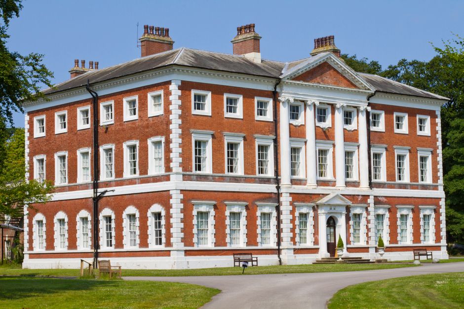 A large, multi-story red brick building with white decorative trims and multiple windows, situated in a landscaped area with grass and trees. The structure features classical architectural elements, including white columns at the entrance and white quoins at the corners. The roof is pitched with several chimneys visible along the top. There are no furniture, boxes, or moving equipment visible inside or around the building in this image, indicating the property is not currently in the process of being moved or packed. However, this image relates to house removals and relocation services offered by Enfield Movers near Forty Hall, ENFIELD, as depicted by the pristine condition and formal appearance of the property, which may be involved in home relocation projects. The overall environment is bright and well-lit with natural daylight highlighting the building's exterior details.