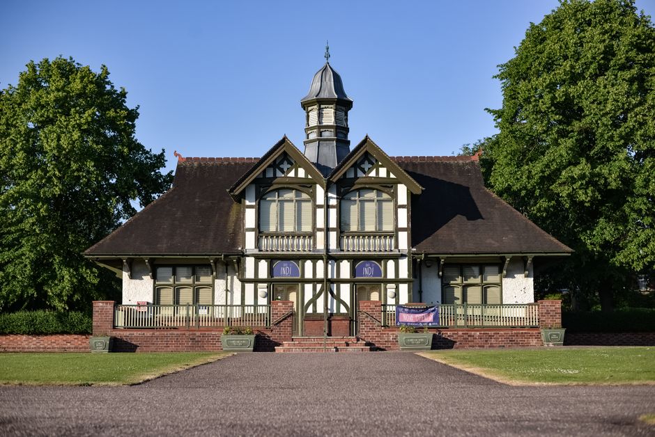 A two-storey historic house situated in a landscaped area with a paved driveway leading up to the entrance. The house features a Tudor-style façade with decorative black wooden beams contrasting with white walls, and a steeply pitched roof with dark shingles. The upper floor has a centered large arched window with a small balcony surrounded by a wooden railing on each side. There are two-folded window shutters on either side of the door, and a small turret with a conical roof rises above the structure. The front entrance has brick steps and a wooden door partially visible behind a small archway. Surrounding the house are two large, leafy green trees on each side, casting shadows on the building, and a well-maintained green lawn edging the pathway. The house appears to be used for community or historical purposes, with banners and signs visible near the front entrance. The overall scene is lit by clear daylight with a bright blue sky, illustrating a home relocation or property transfer context typical in house removals or moving services, as managed by Enfield Movers.