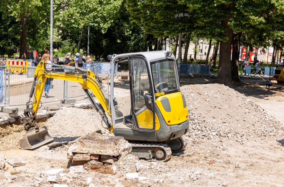 A compact excavator with a white cab and red base is positioned in a construction site in front of a multi-storey building with dark brown brick exterior walls and white-framed windows. The excavator'